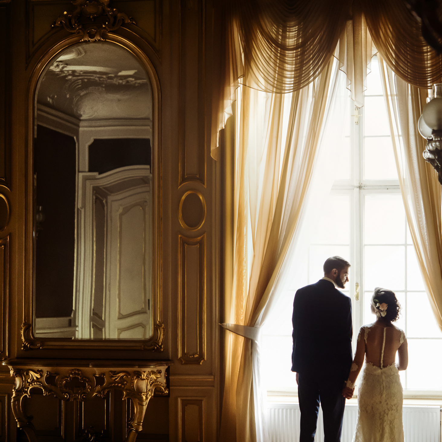 stylish luxury bride and handsome elegant groom holding hands at gorgeous window on the background of rich interior in old building